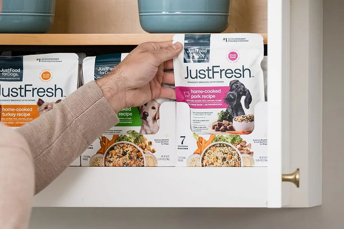A hand pulls a JustFresh shelf-stable pouch from a kitchen pantry stocked with JustFoodForDogs meals