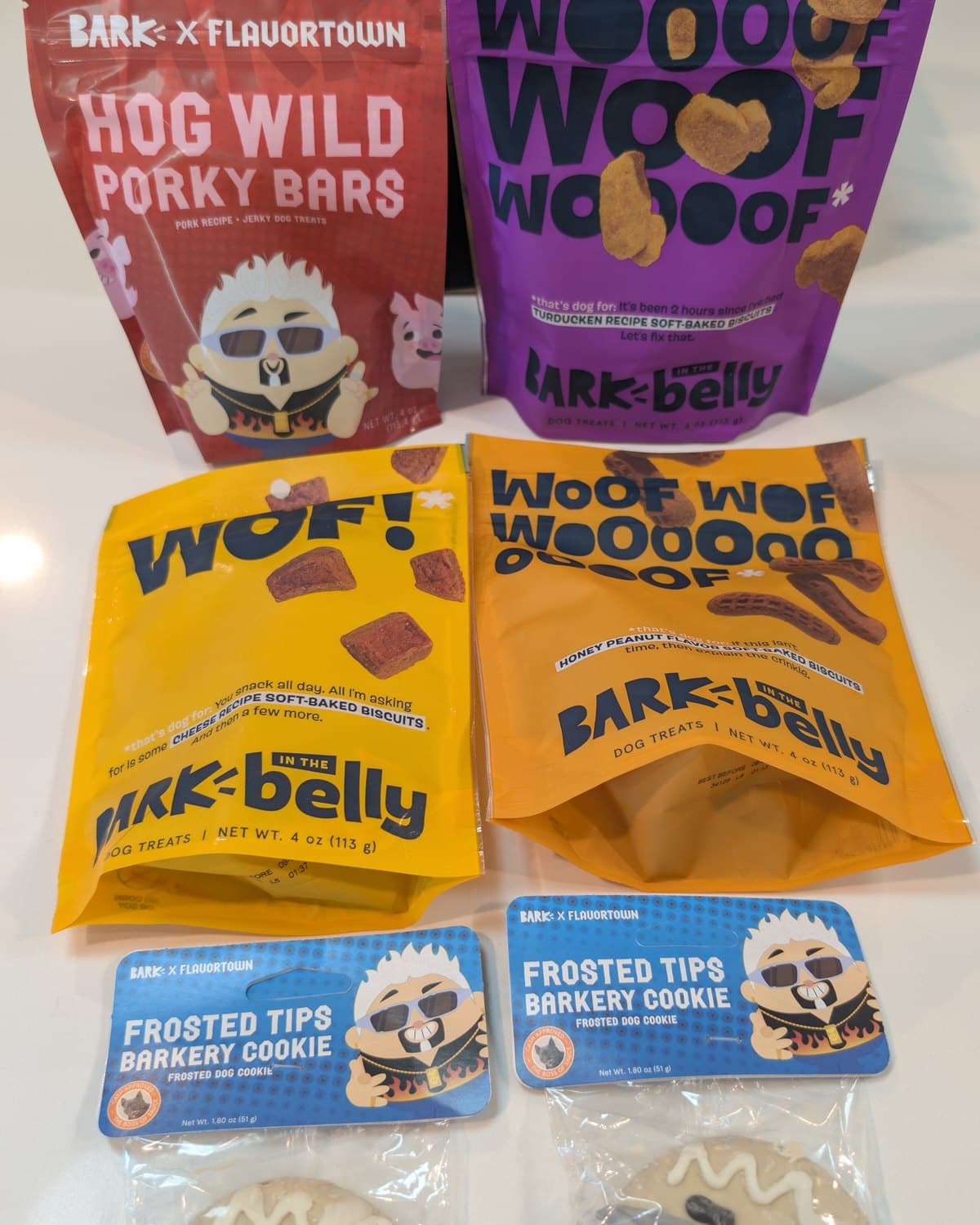 Four BARK treat pouches and two Frosted Tips Barkery Cookies arranged in front of the box