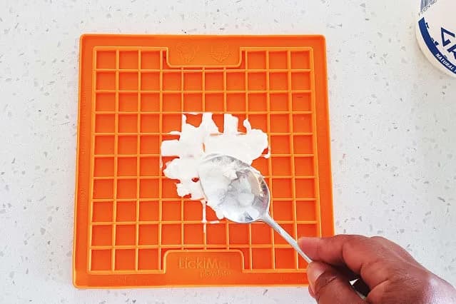 Hand spreading soft food with a spoon on an orange LickiMat Playdate
