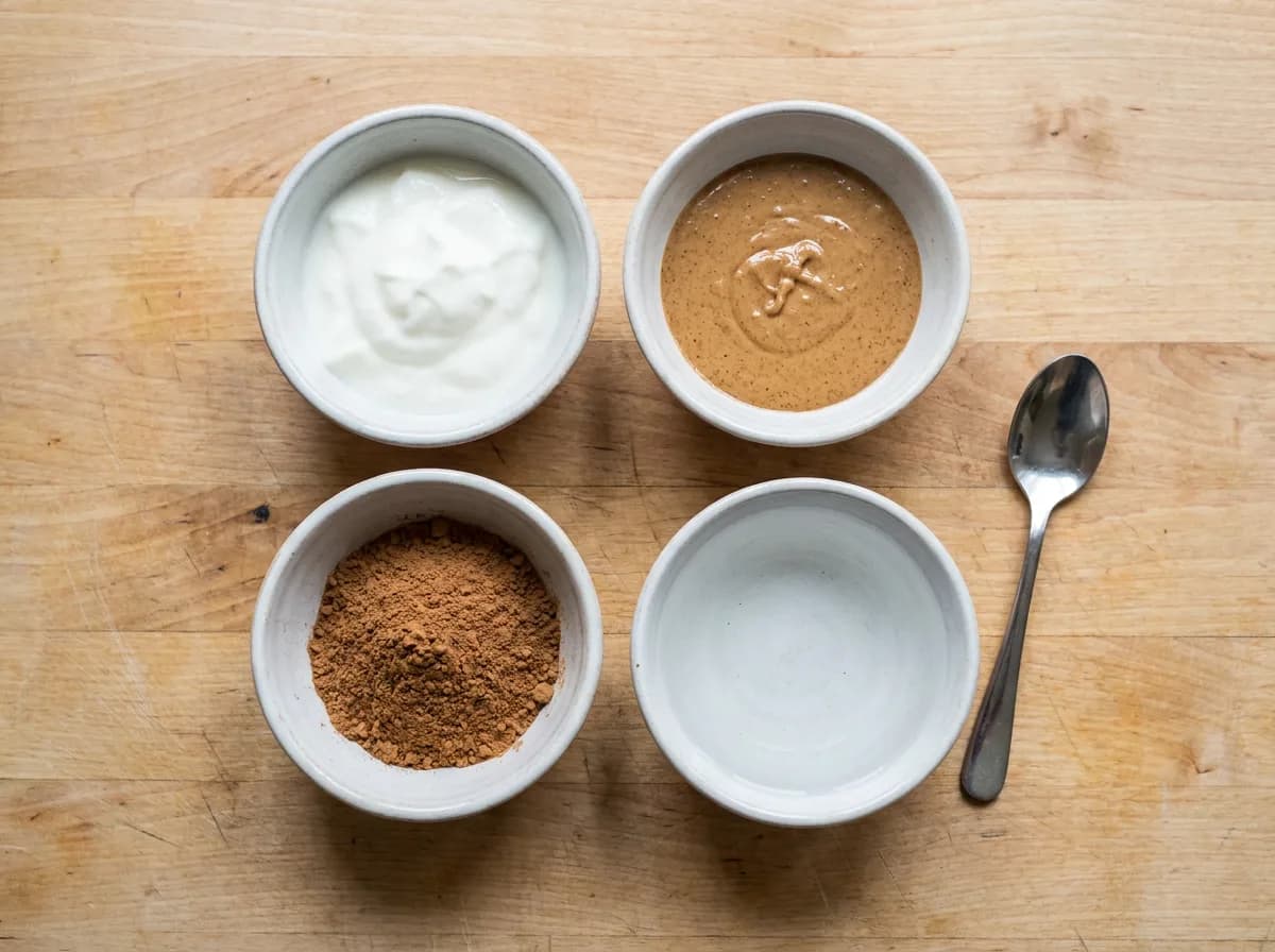 Separate bowls of plain yogurt, almond butter, carob powder and water beside a spoon