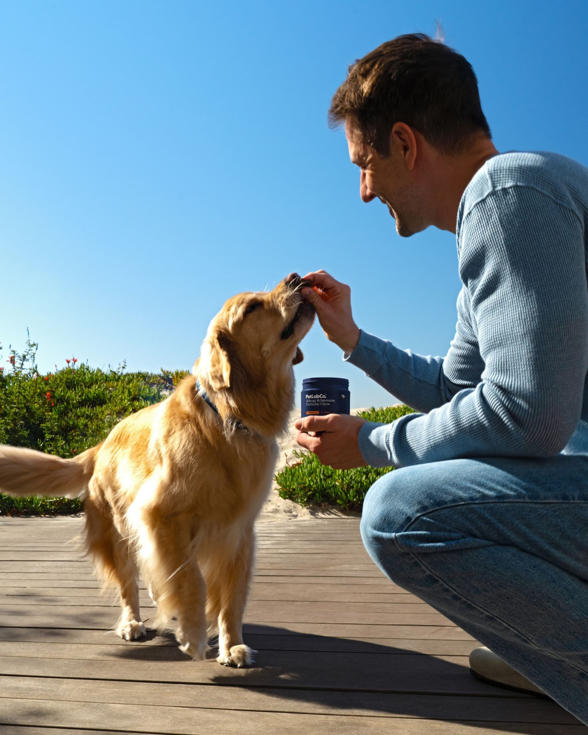 A man holding an Allergy & Immune tub offers a chew to a Golden Retriever outdoors.