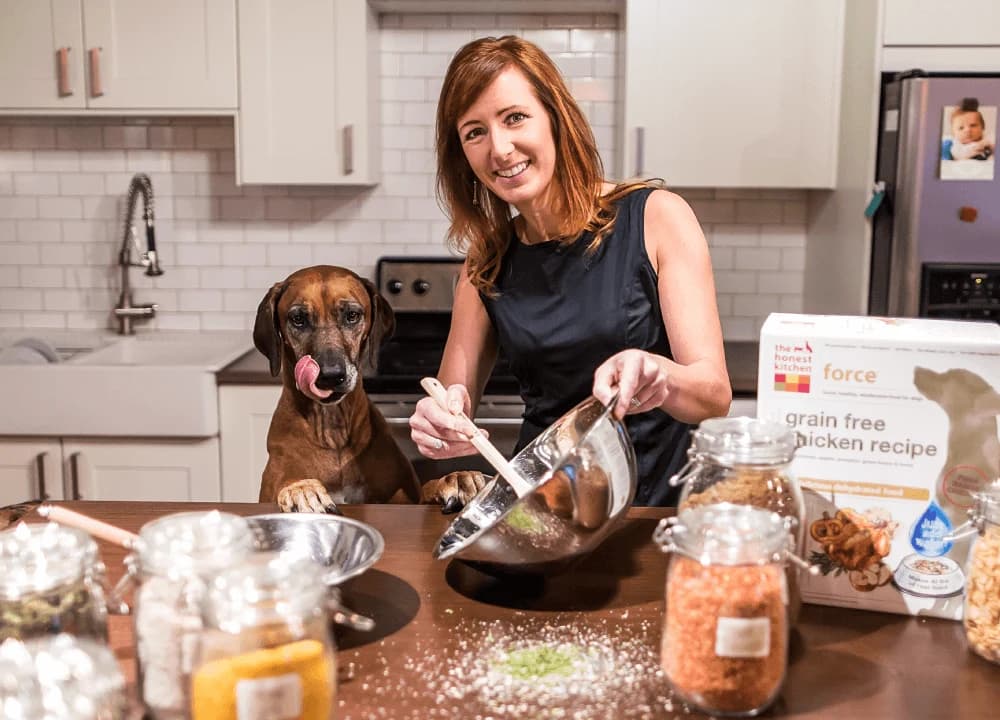 Honest Kitchen founder Lucy Postins preparing food in a kitchen with her dog nearby